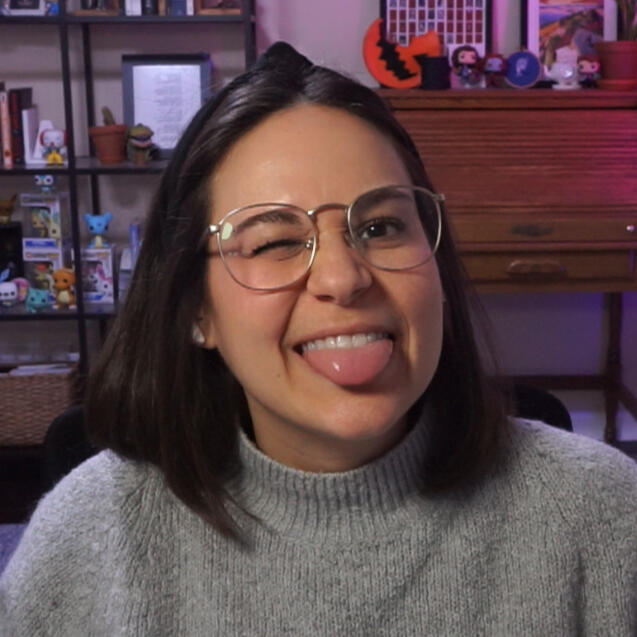 Jamie in glasses & a gray sweater, winking & sticking out her tongue. She has shoulder-length brown hair. In the bg, theres a wooden desk w a roll-top & various trinkets, plants & figurines. To the lefts a shelving unit w books, plants, & more trinkets.