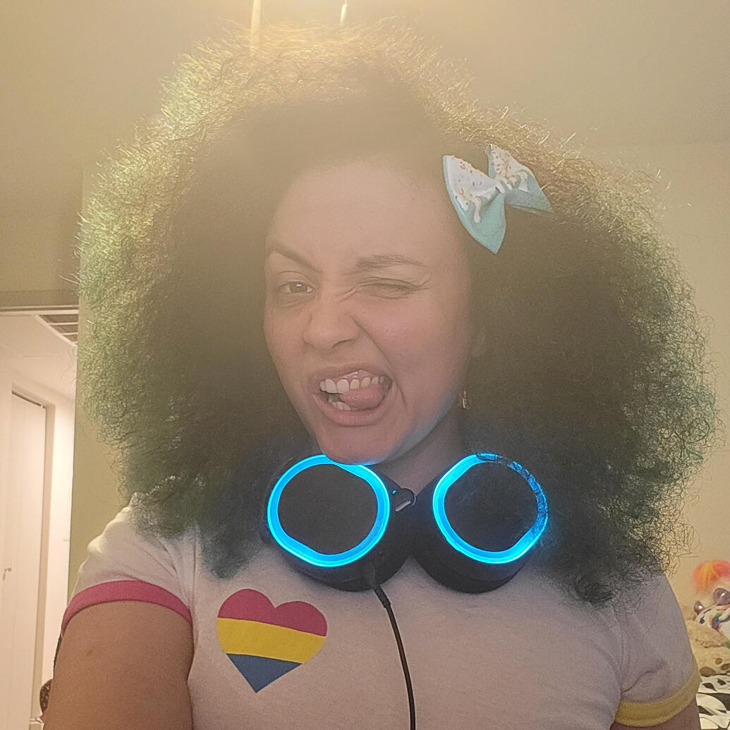 A person winking at the camera with a curly afro and bow. They are wearing a white pansexual themed t-shirt, and blue headphones around their neck.
