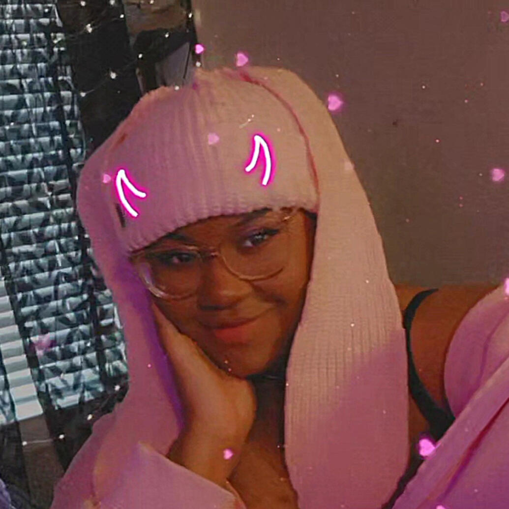 image showing theperfectpxl, a Black content creator wearing clear glasses, a pink beanie with bunny ears, with a hand on her face and a closed lipped smiling wearing a pink hoodie
