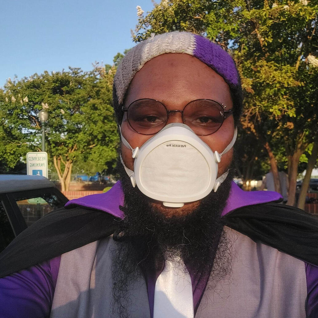 Black individual with a grey vest, white tie and ace colored beanie. Individual is bespectacled and bearded with a background of trees.